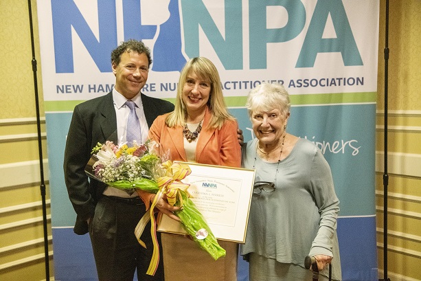 Christine Ferrer holds her floral bouquet and framed award in front of a conference banner, between her husband and mother. 